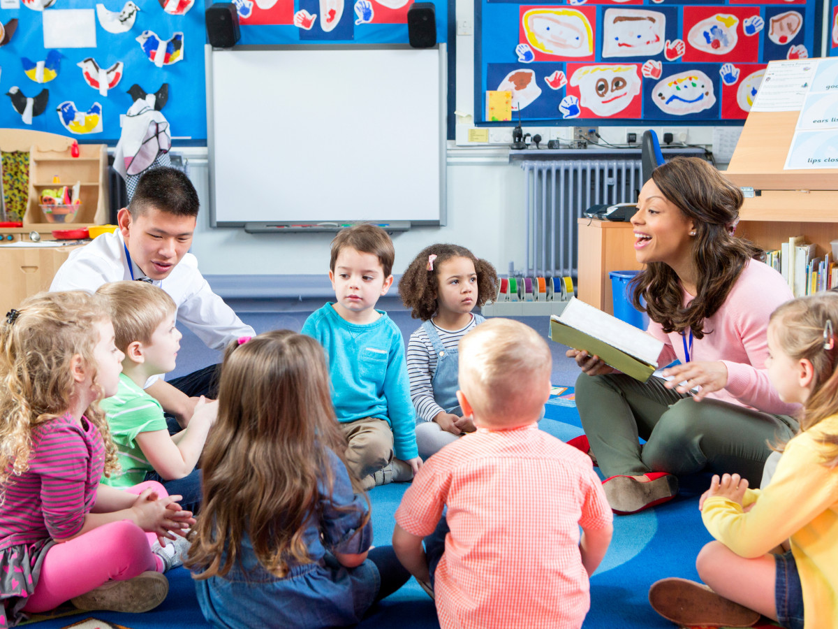 Group of nursery children sitting on the floor in their classroom with their teachers. The female teacher is reading from a book.